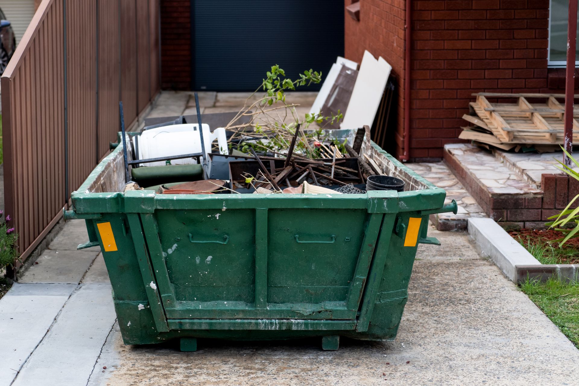 Green dumpster filled with construction debris and yard waste in a driveway near a house