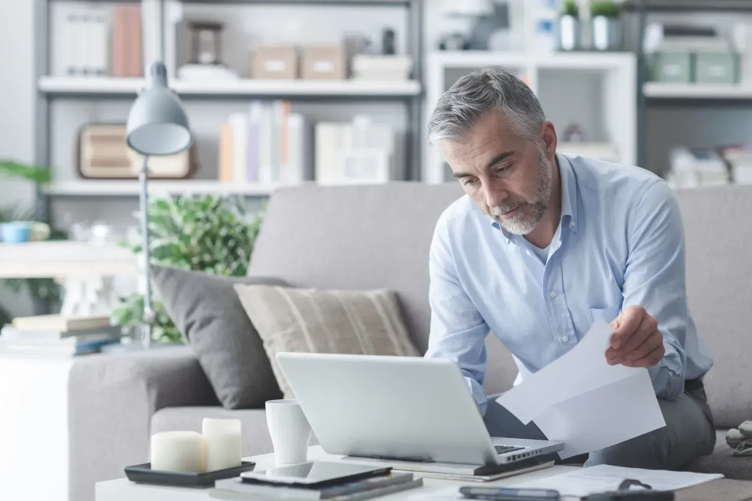 Man sitting on couch, working on laptop, reviewing paperwork. Interior home setting.