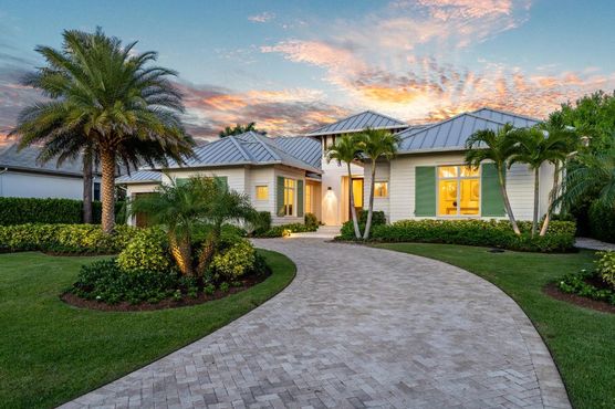 Modern two-story house with stone facade, palm trees, and illuminated windows at dusk.