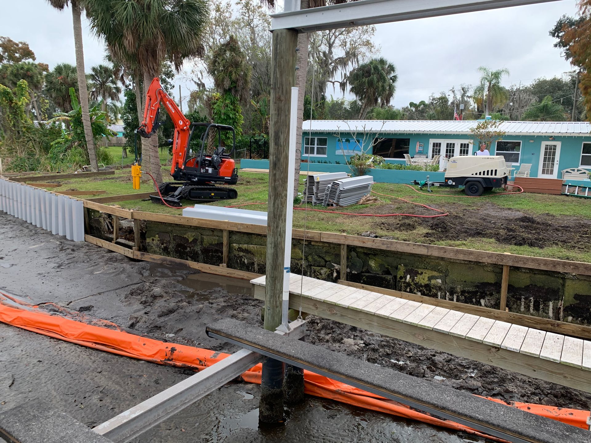 Construction site with an excavator near a wooden bulkhead, residential house in the background.