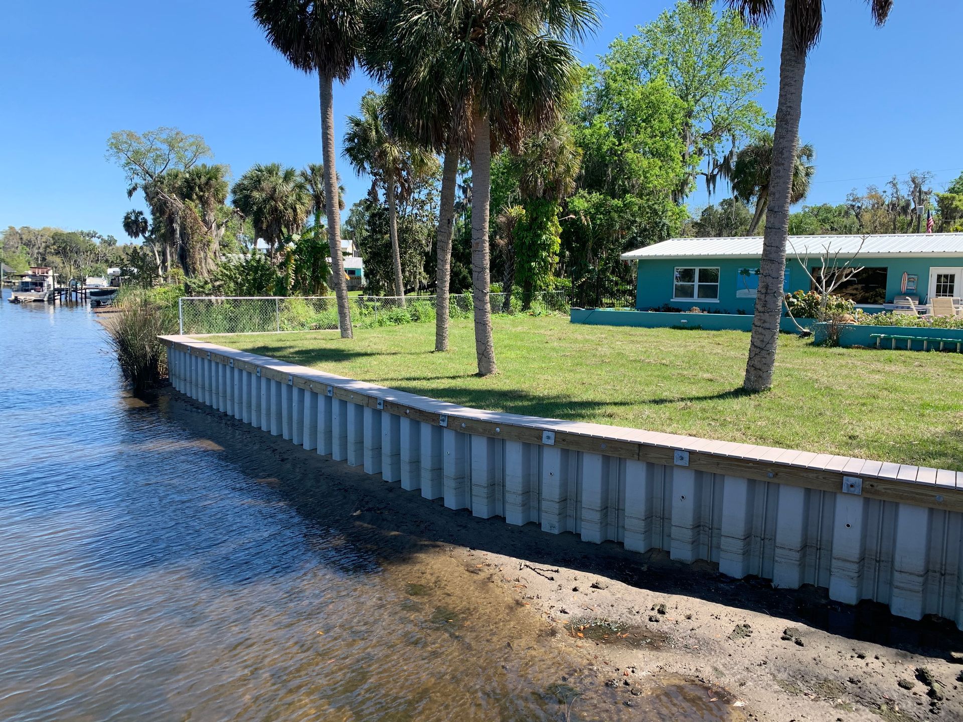Waterfront with a seawall and a light blue house under a bright blue sky. Palm trees and green grass.