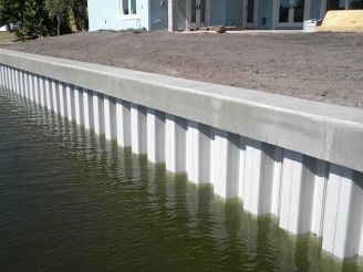 A white and gray seawall bordering water, with a concrete top, next to a building and dirt.