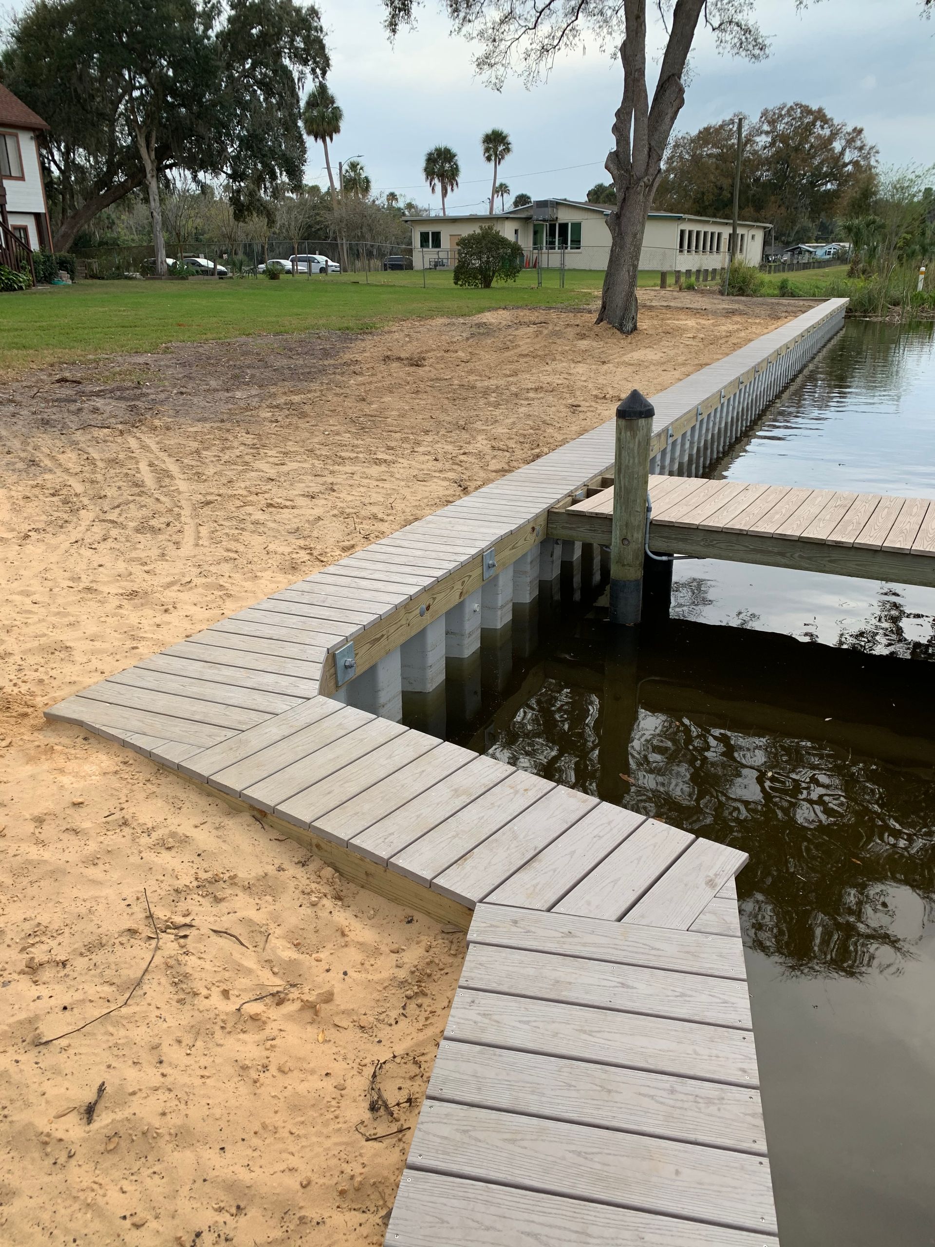 Wooden boardwalk leading to a dock by a shoreline with sand and water, next to buildings and trees.