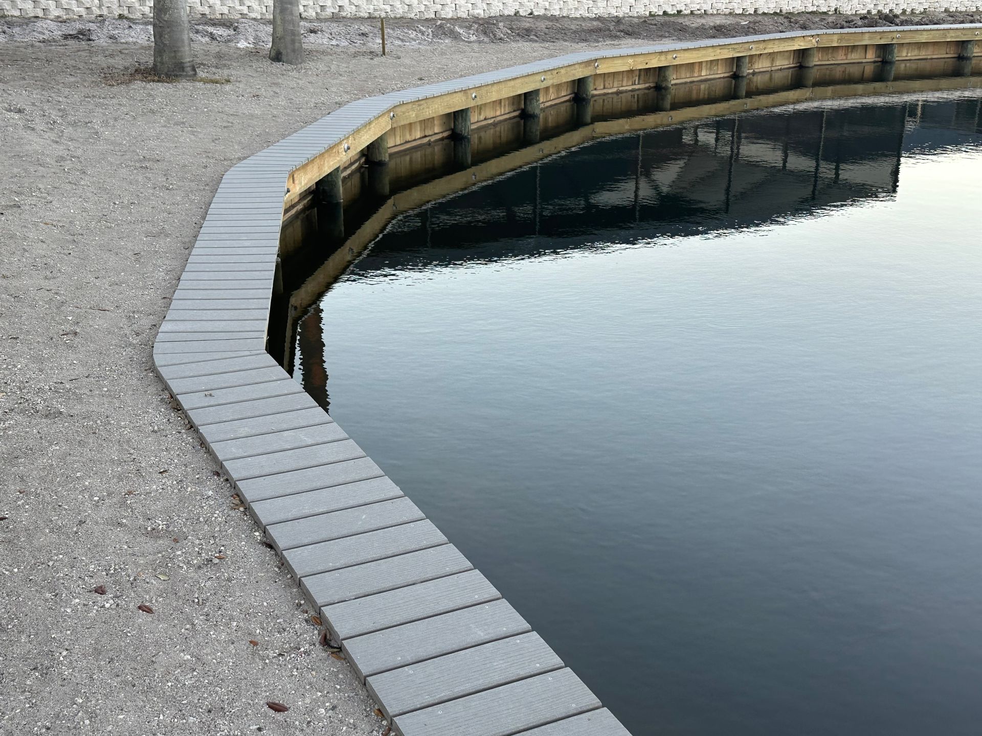 Curved shoreline with gray composite edging, reflecting water, gravel path, and wood support beams.