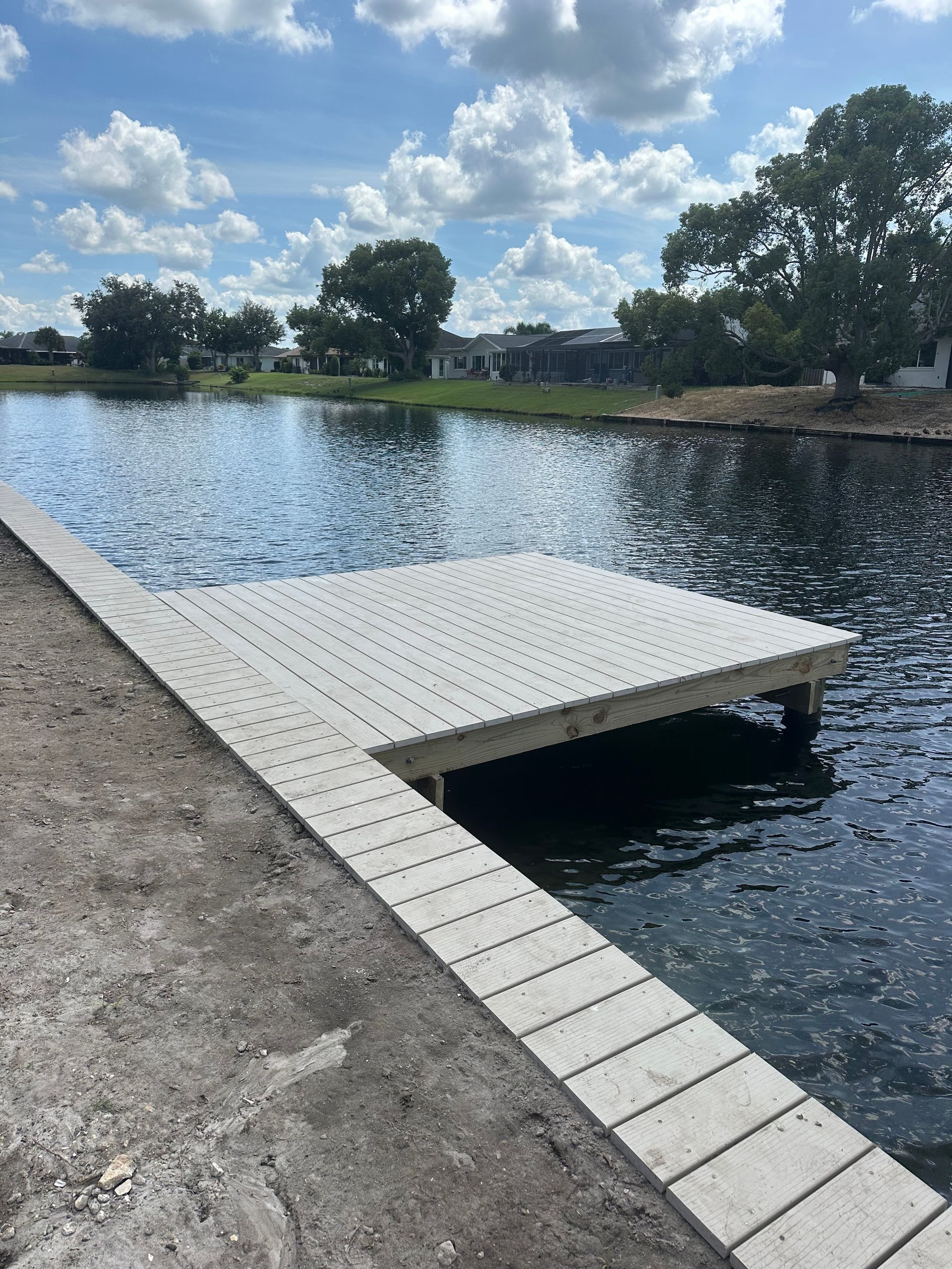 Dock extending into a lake, gray plank surface. Blue sky, water, and trees in background.