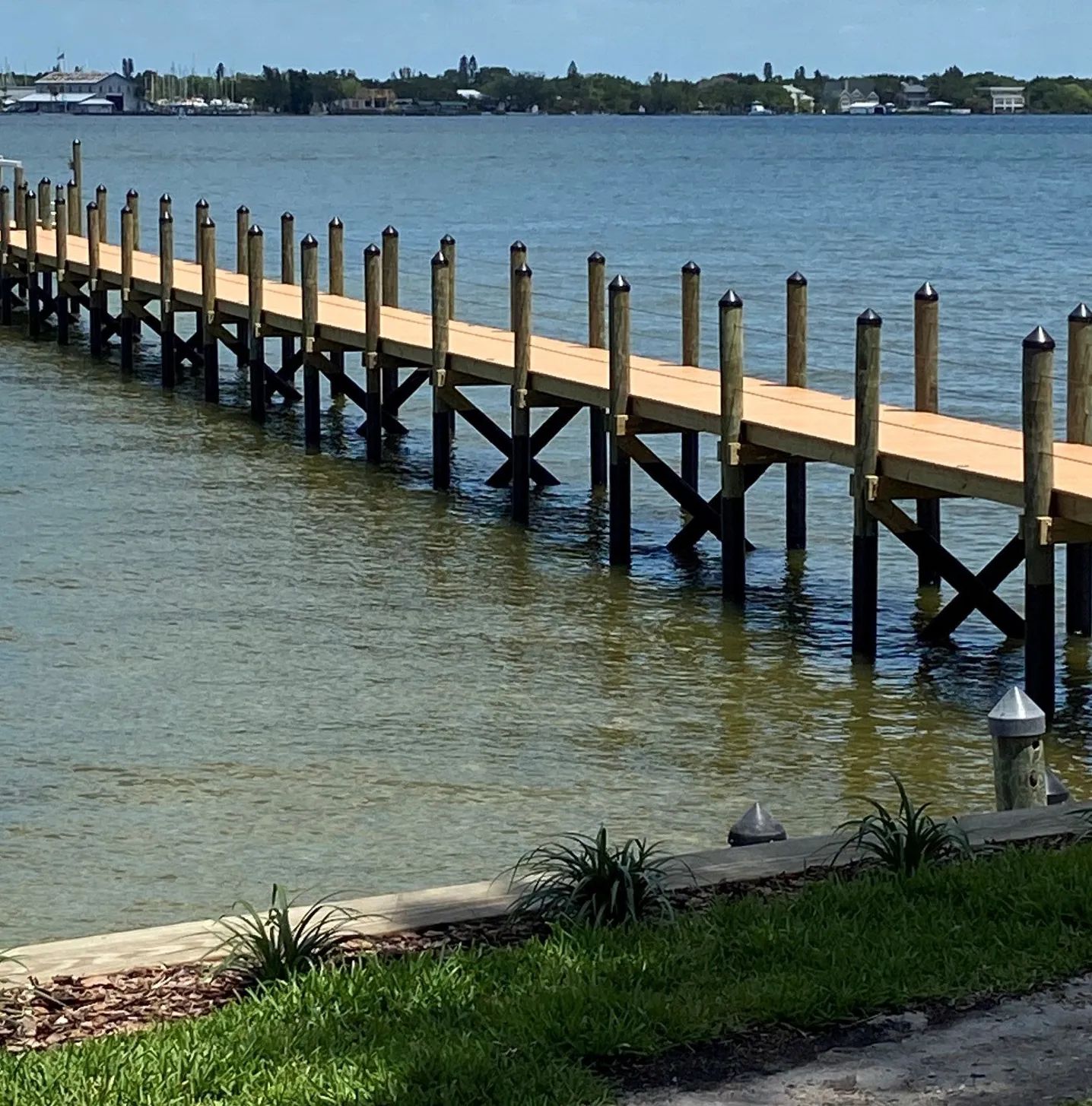 Wooden pier extends into calm blue water. Green grass and distant buildings in the background.
