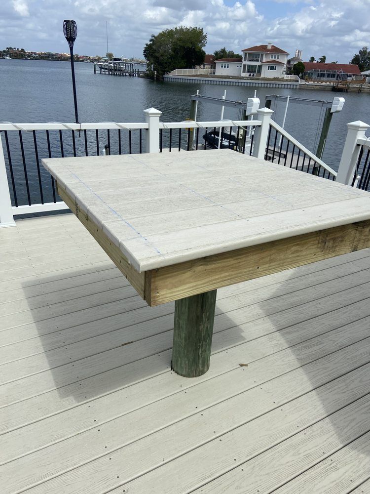 Square concrete-topped table on a wooden deck overlooking a waterway, with a house and boats in the background.