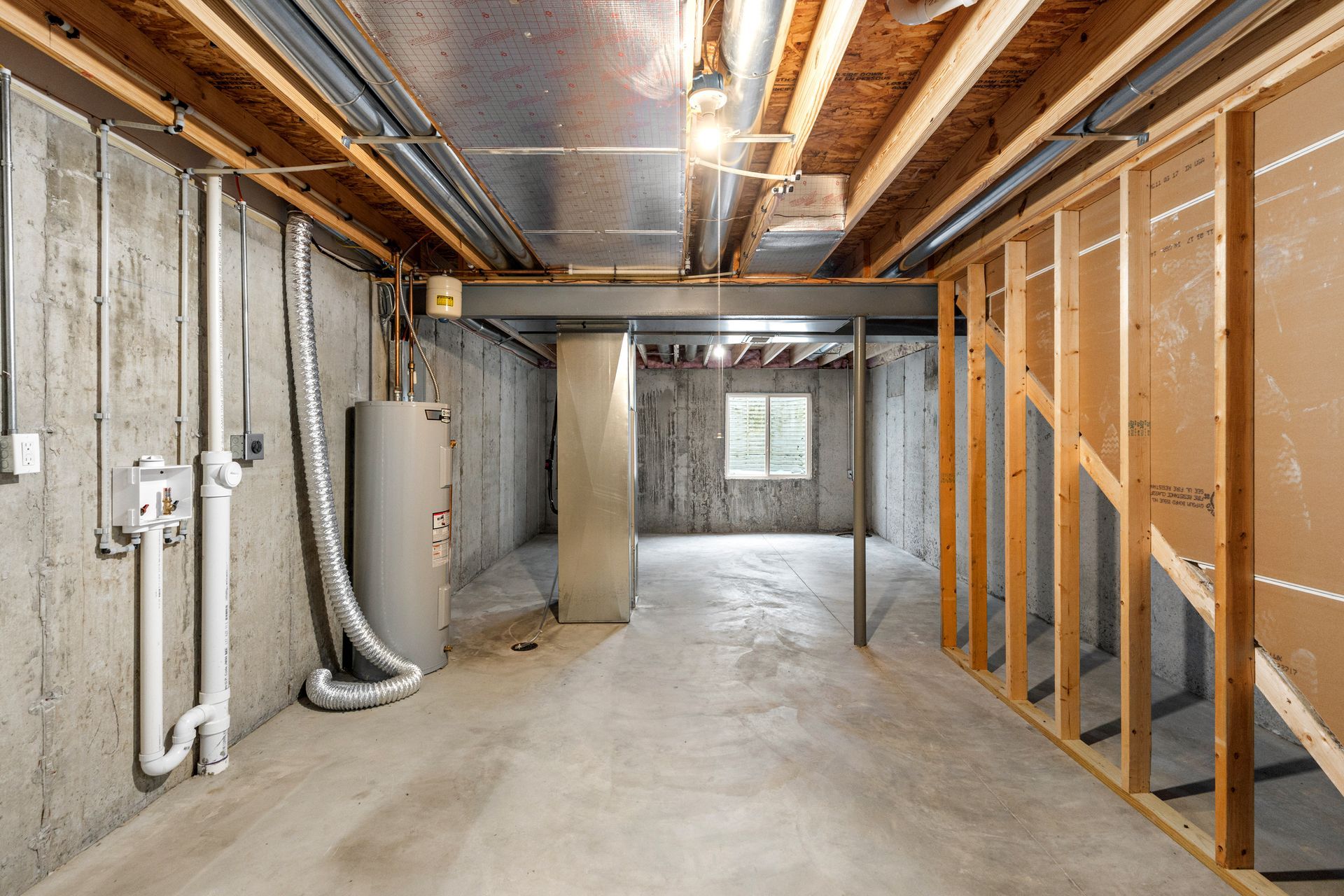 Unfinished basement with exposed beams, pipes, insulation, and a concrete floor opening into a room with a small window.