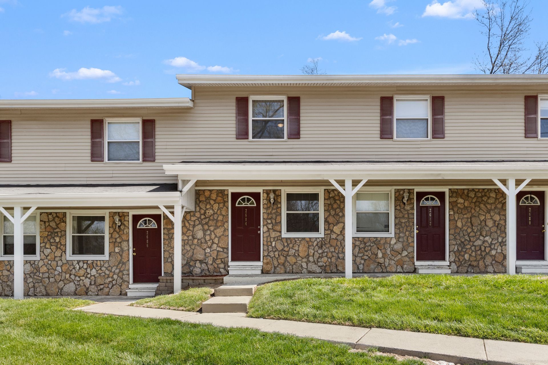 Two-story townhouse row with brick facades, white trim, maroon doors, and a green front lawn under a blue sky
