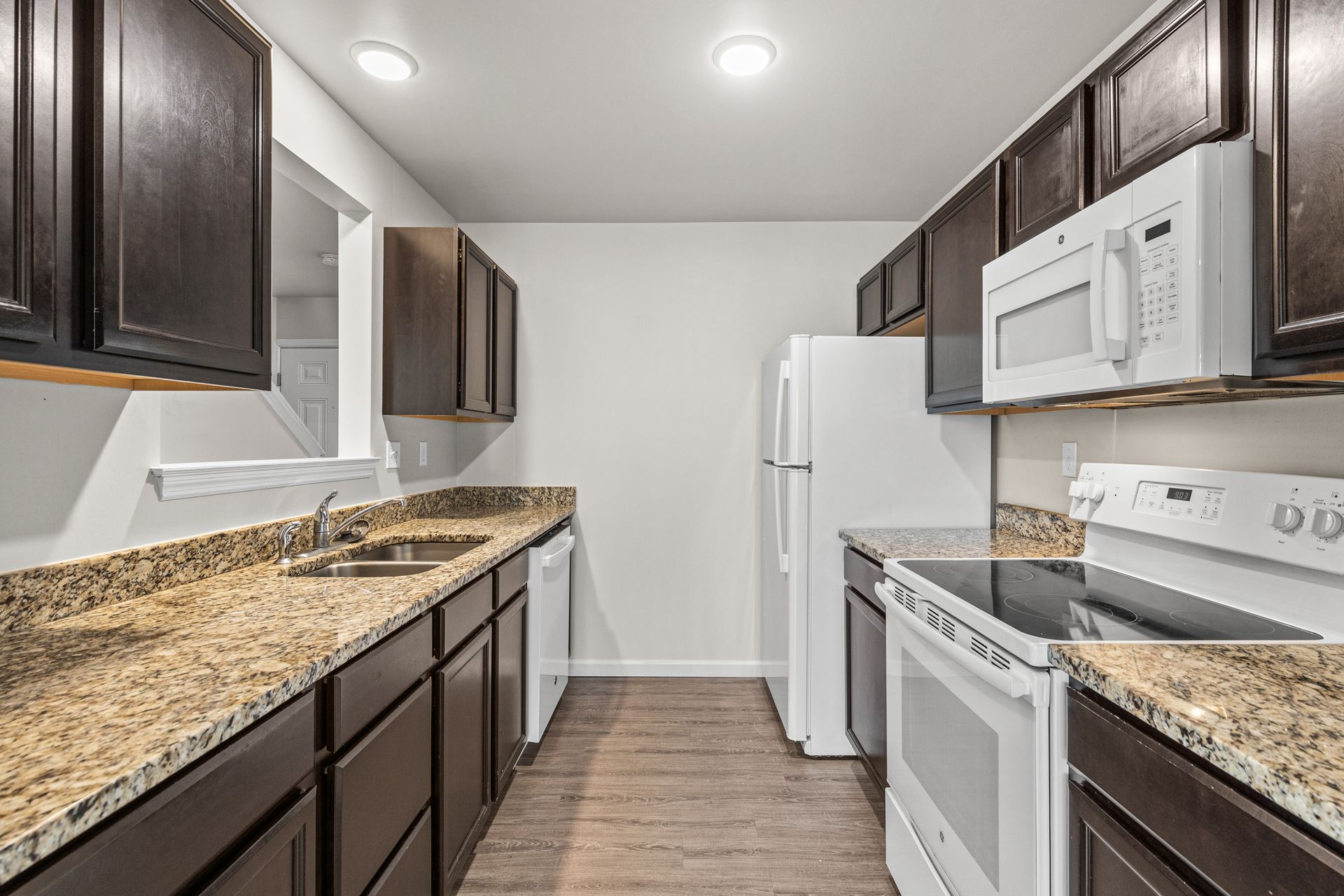 Modern galley kitchen with dark wood cabinets, speckled countertops, white appliances, and wood-look flooring
