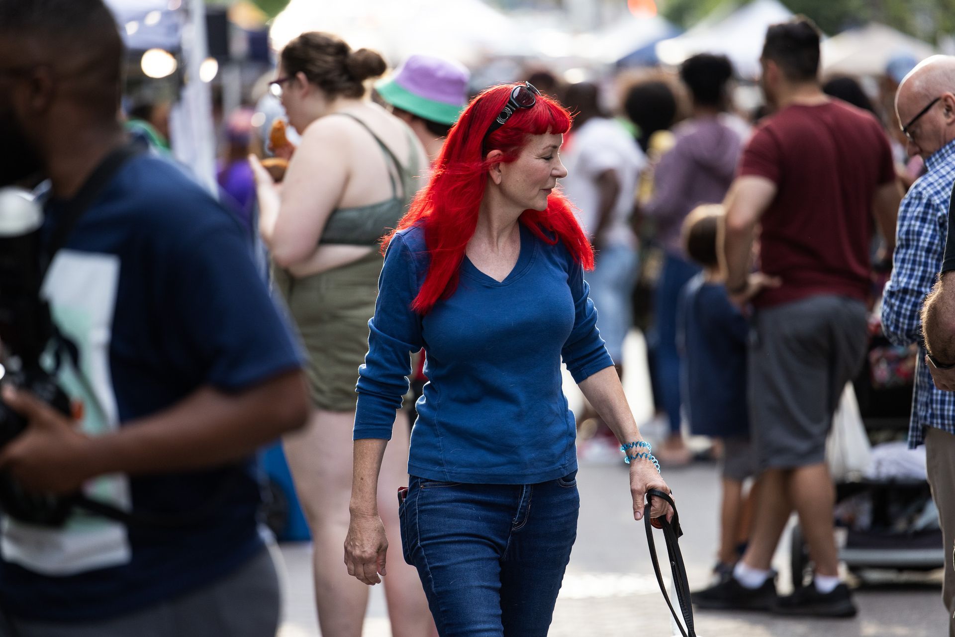 Woman with bright red hair walking through a crowded street, holding a bag and wearing a blue top and jeans