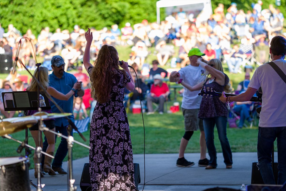 Outdoor concert performance on a stage, with musicians and dancers in front of a seated crowd.