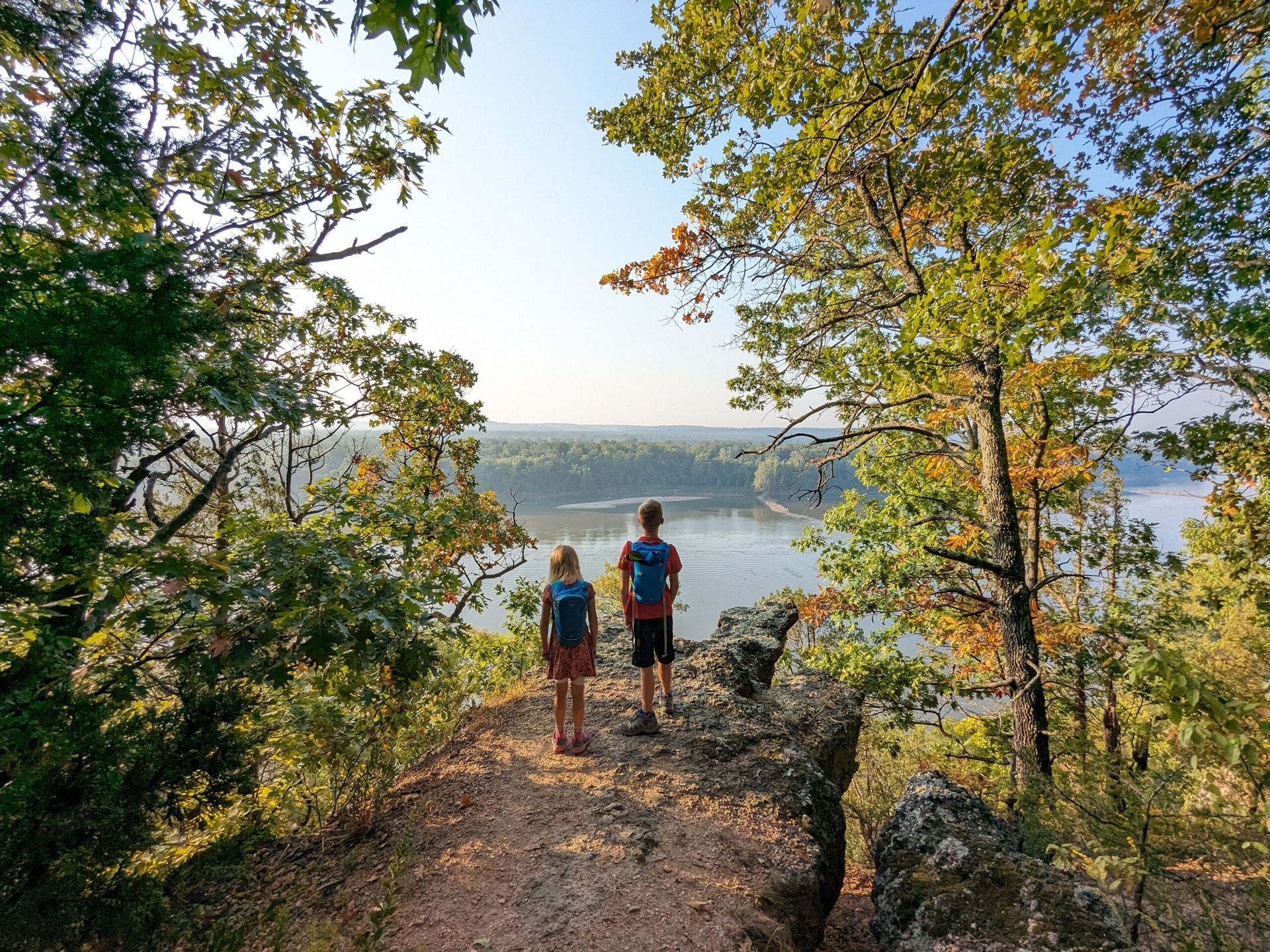 Two hikers on a forest trail overlook a calm lake under leafy trees