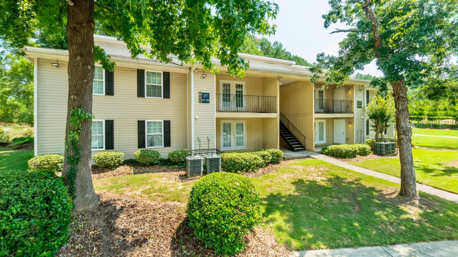 Apartment complex with tan buildings, green lawns, and a paved parking lot under a sunny sky