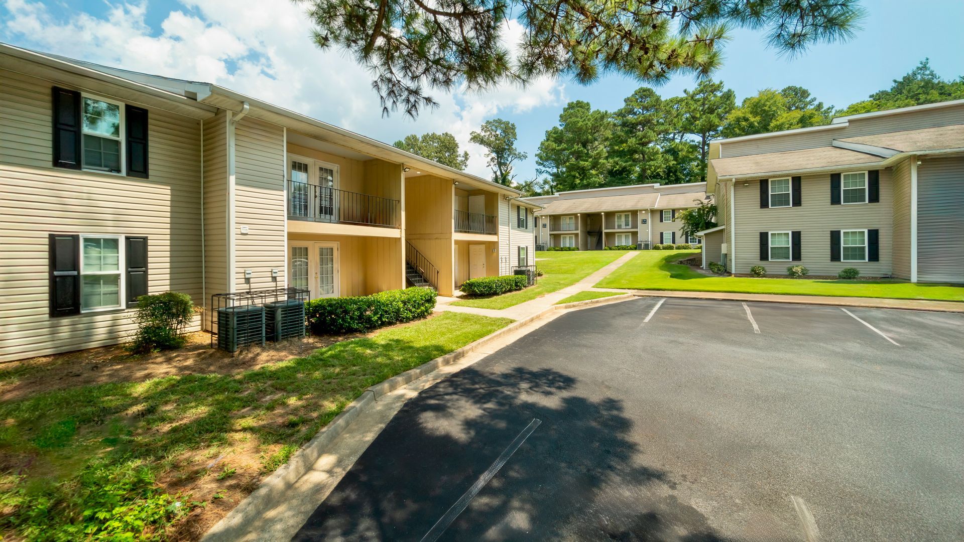 Apartment complex courtyard with beige buildings, green lawns, and a paved parking lot under a sunny sky