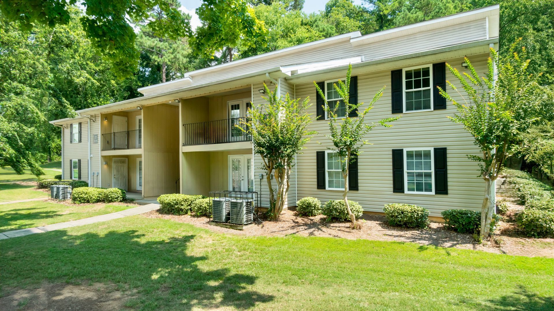 Apartment complex driveway with beige buildings, green lawn, and trees under a blue sky