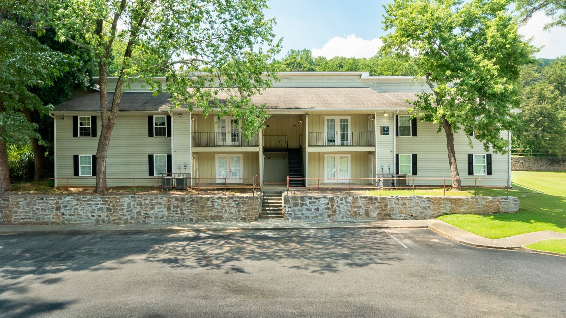 Two-story beige apartment building with stone retaining wall and trees in front