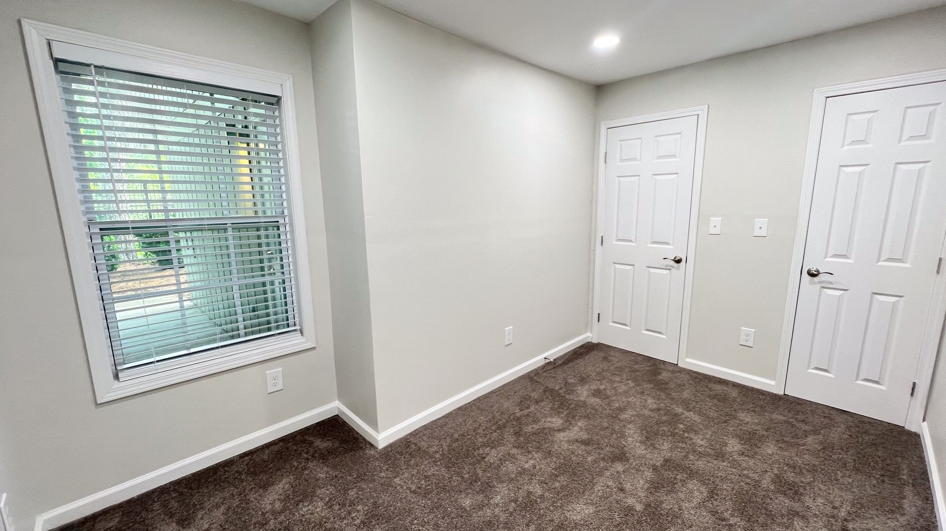 Empty beige bedroom with carpet, window blinds, recessed light, and two white closet doors