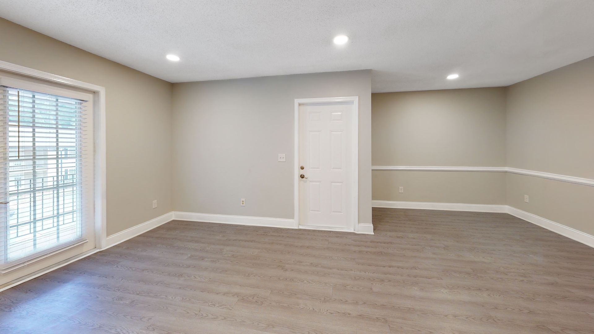 Empty beige living room with gray wood flooring, white trim, and sliding glass doors on the left