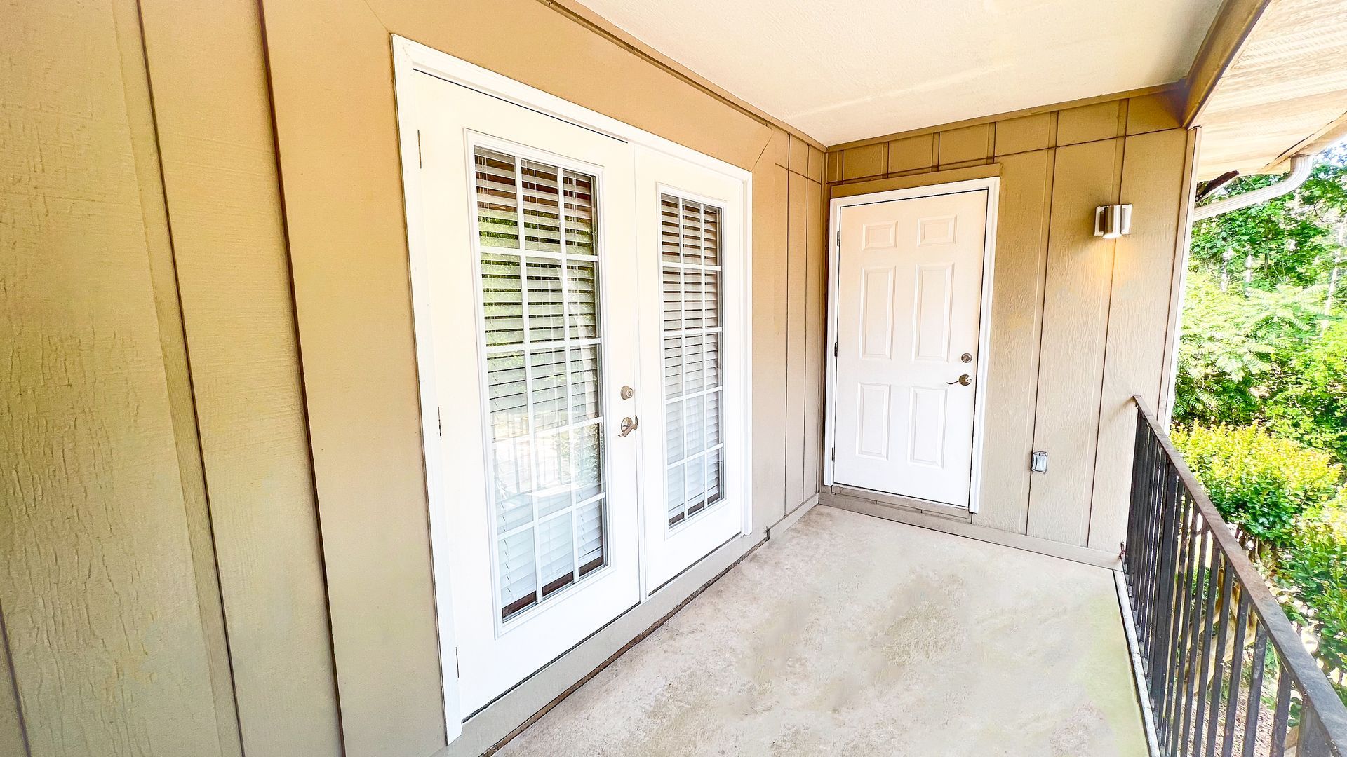 Covered apartment balcony with beige walls, white French doors, and a railing overlooking greenery