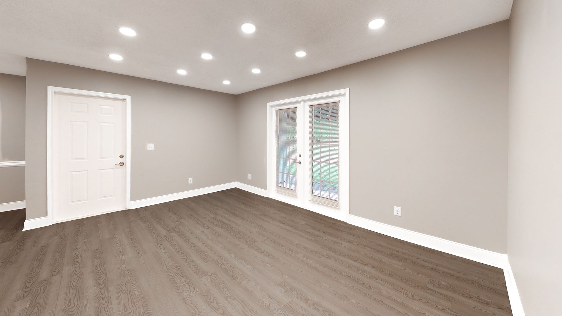 Empty modern living room with gray walls, wood flooring, recessed lights, and a windowed door.