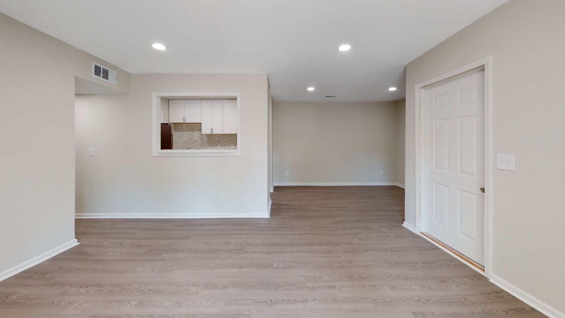Empty modern living room with gray wood floors, white walls, recessed lights, and a pass-through opening to the kitchen.