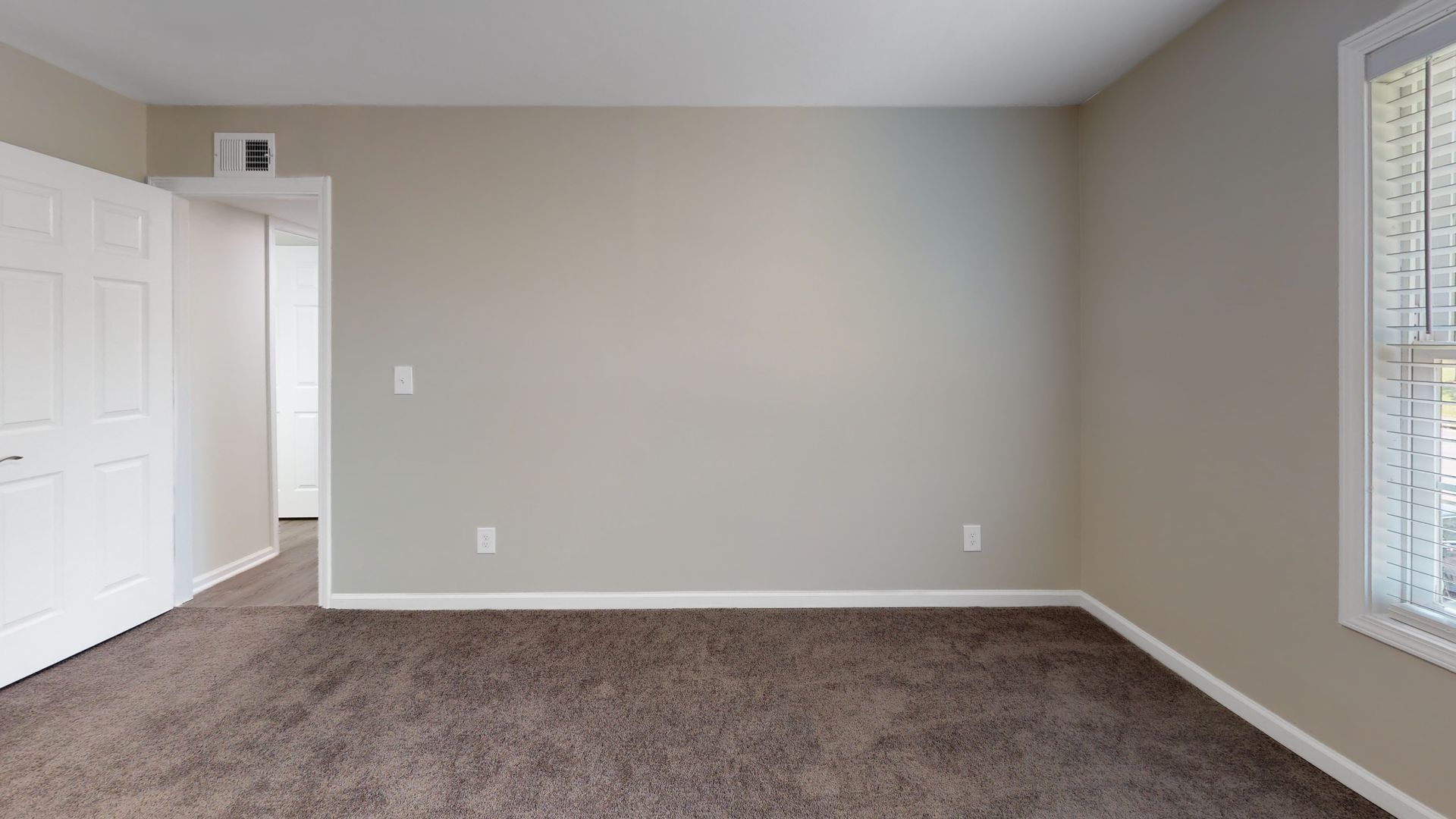 Empty beige carpeted room with white walls, closet doors, and a window with blinds