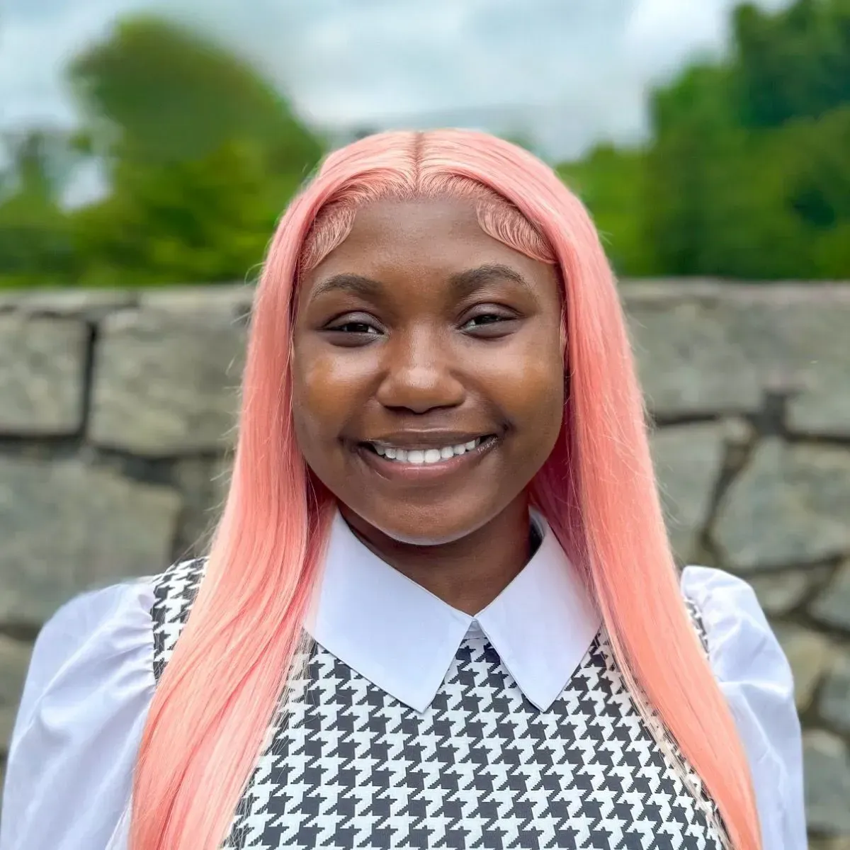 Smiling person with pink hair wearing a white shirt and houndstooth vest outdoors in front of a stone wall.