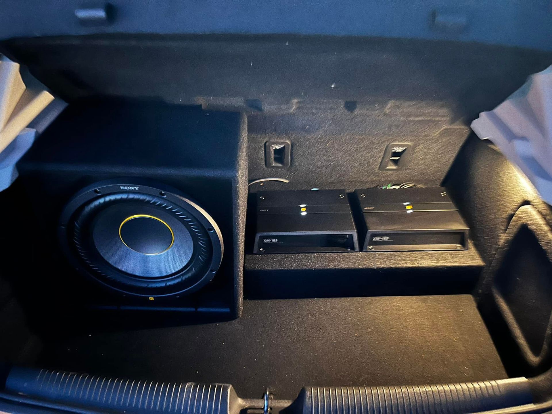 Car trunk with a large subwoofer speaker on the left and folded rear seats with power outlets in back.