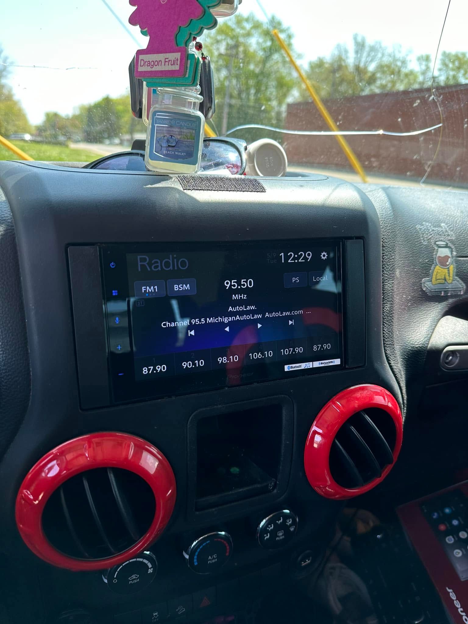 Car dashboard with center touchscreen, red air vents, and hanging air freshener near the windshield.