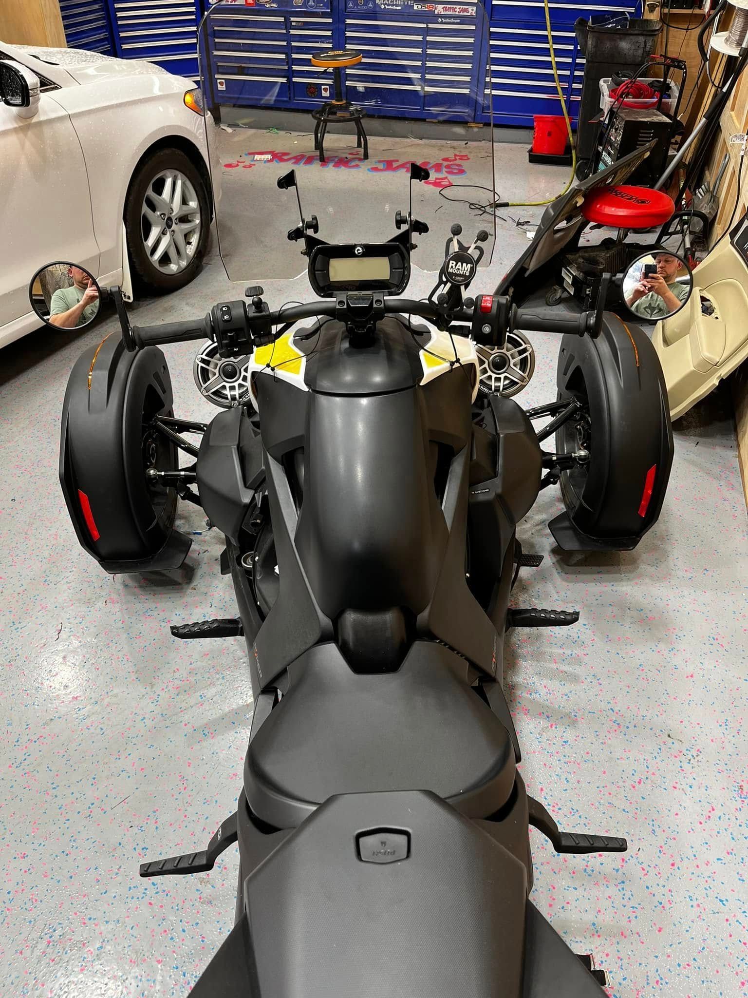 Three-wheeled matte black motorcycle in a garage, viewed from the rear with other cars nearby.