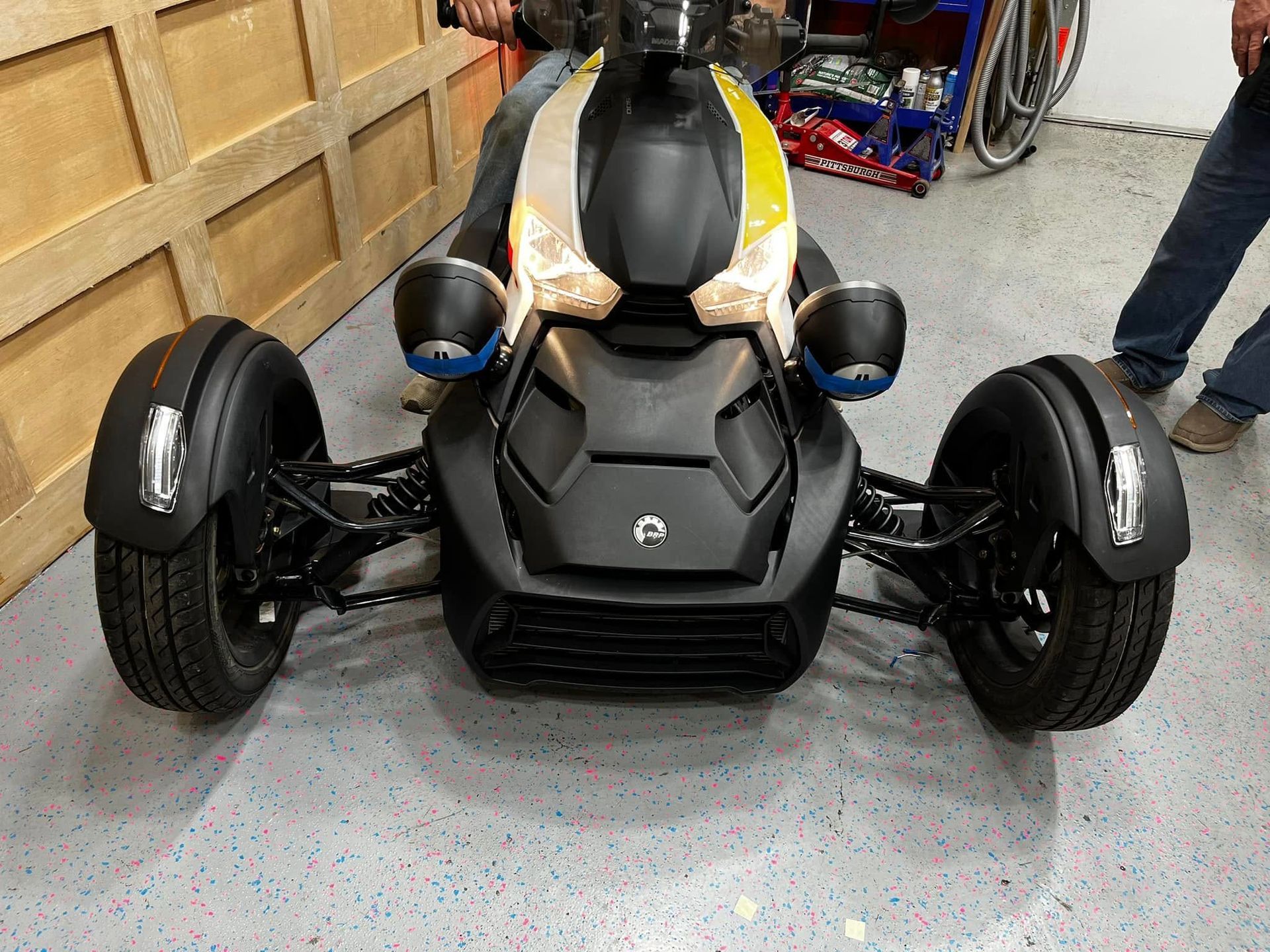 Black three-wheeled roadster with blue mirrors in a garage, front view