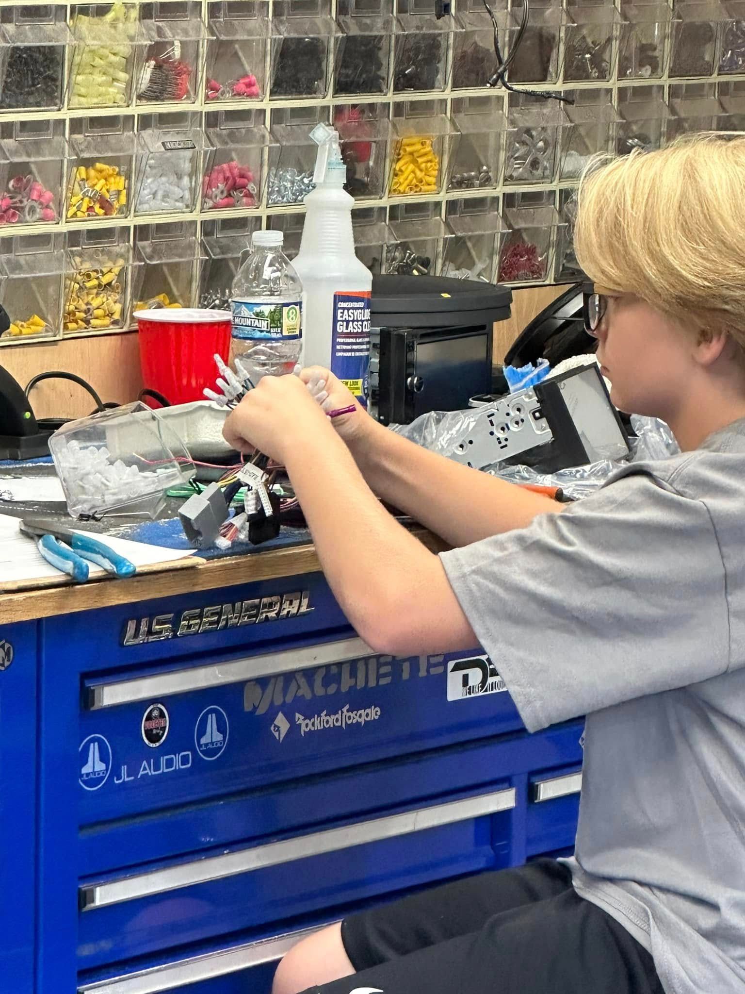 Person working at a blue toolbench in a workshop, handling parts near a vise and organized storage bins.