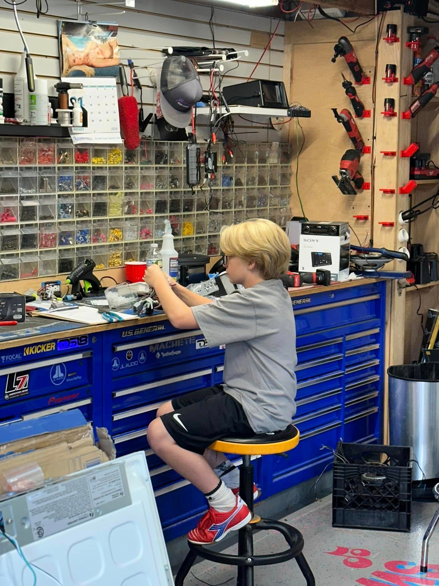 Person sitting on a stool in a workshop, working at a cluttered blue toolbench.