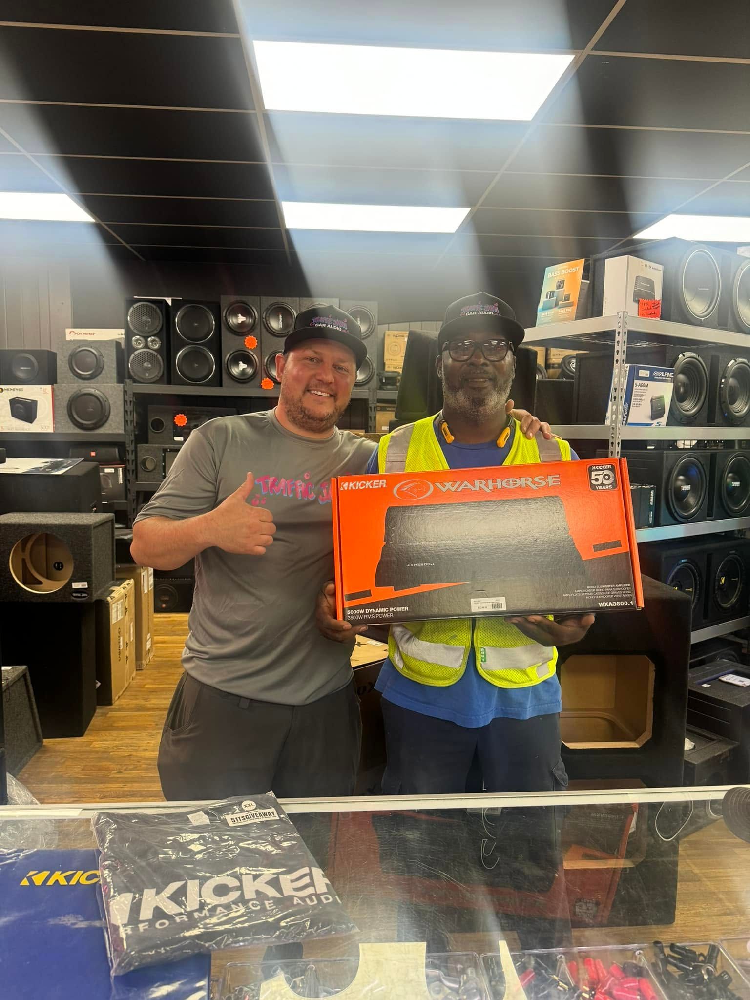 Two people in a store holding a large orange box, smiling among stacked speakers and electronics.