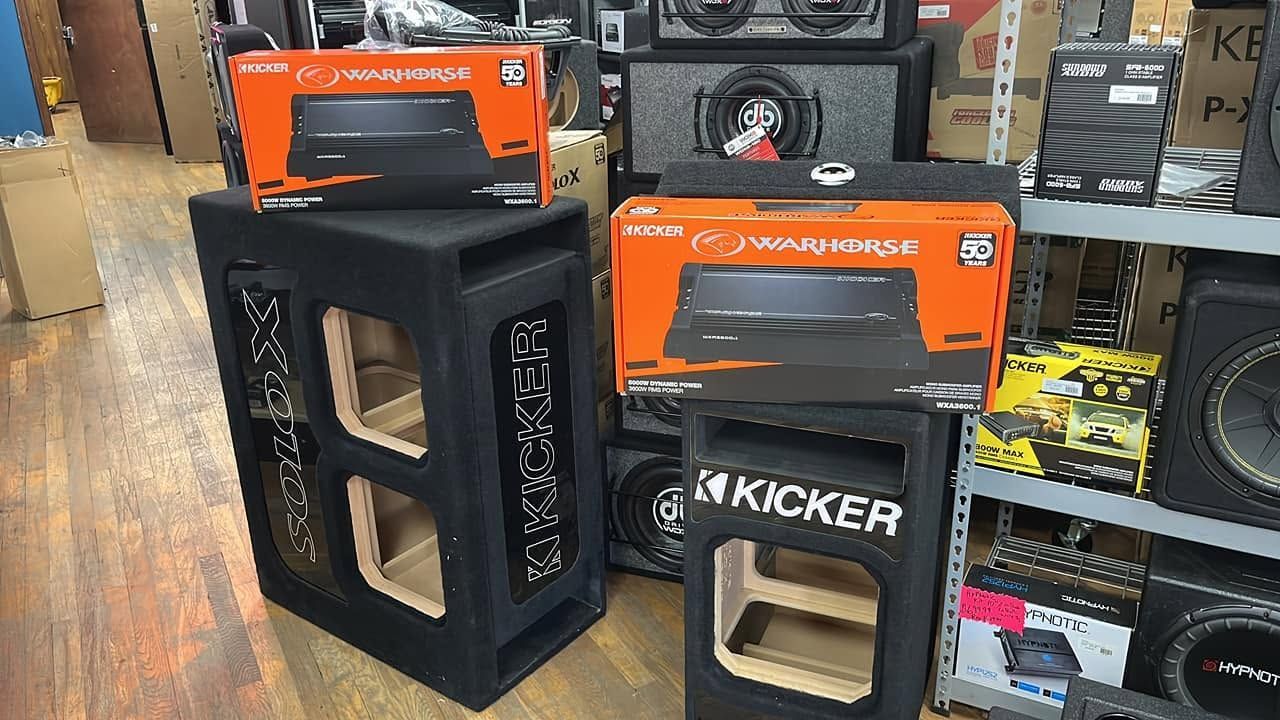Kicker car audio subwoofer boxes stacked in a shop aisle, with orange product packaging behind them