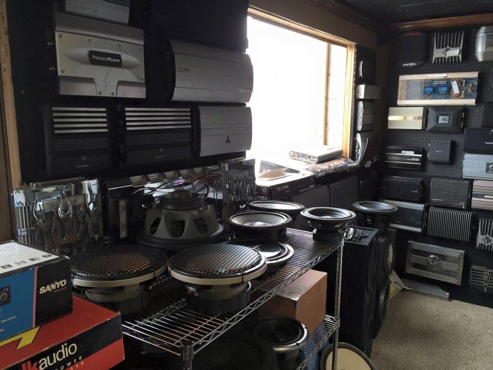 Room filled with stacked audio equipment, speaker parts, and boxes beside a bright window