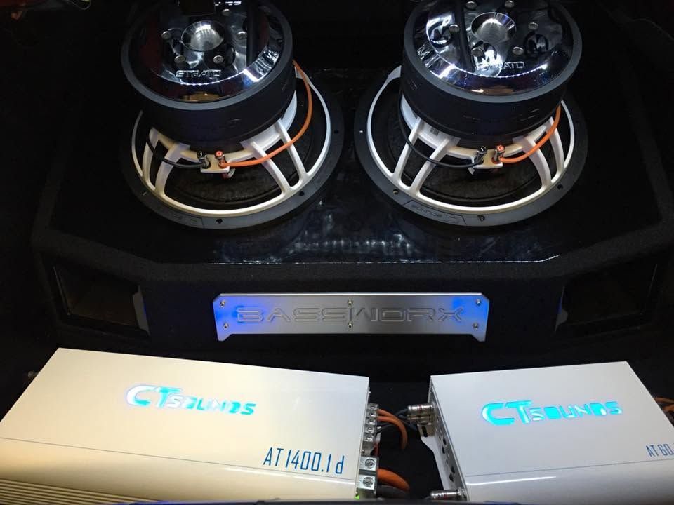 Car audio setup with two large subwoofers and amplifiers in a black enclosure, lit by blue LEDs