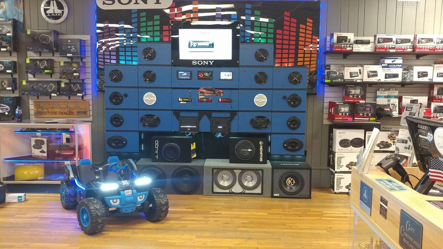 Blue RC truck with bright headlights displayed in a hobby shop in front of stacked speakers and accessories