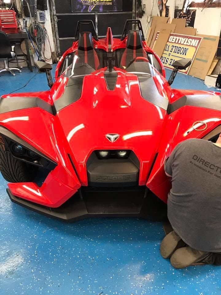 Red three-wheeled sports car in a garage, front view with black accents and a person kneeling beside it