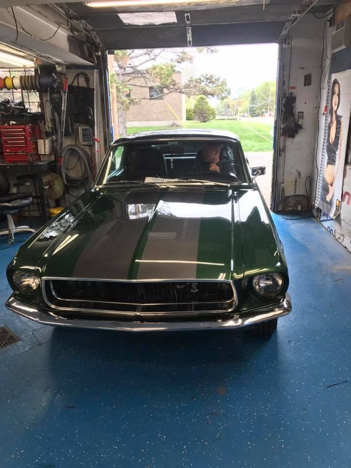 Green classic Mustang with white racing stripes parked in a garage with a blue floor.