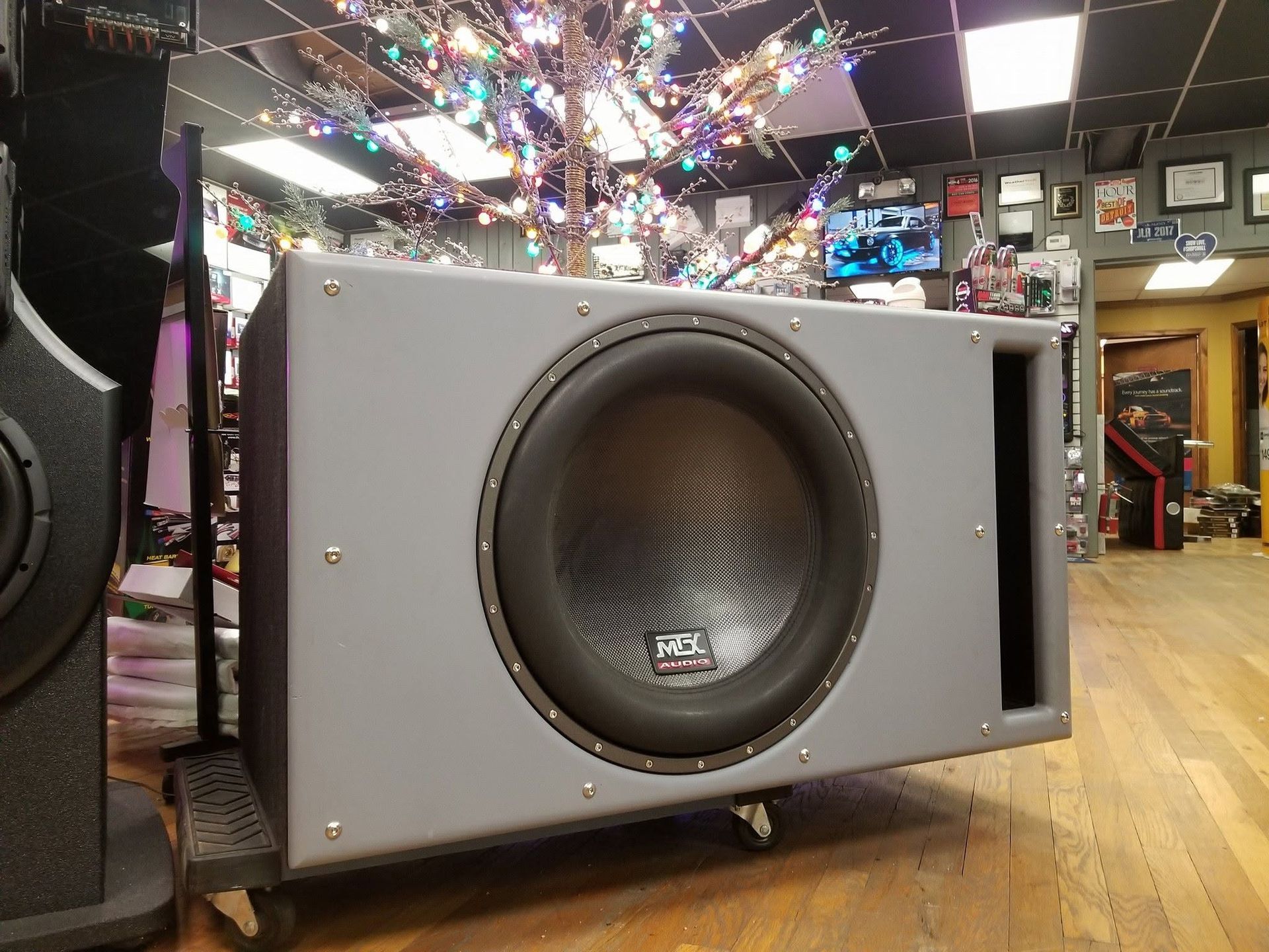 Gray subwoofer speaker on a wood floor in a music store, with colorful lights and other equipment behind it