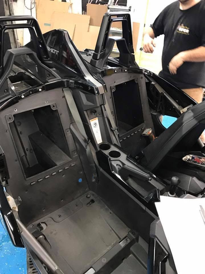 Black vehicle chassis with two seats and open frame, shown in a workshop with people nearby
