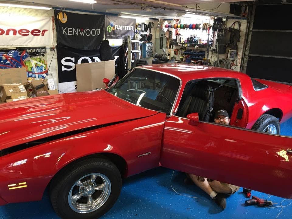 Red classic sports car in a garage, with a person visible inside and the driver’s door open.