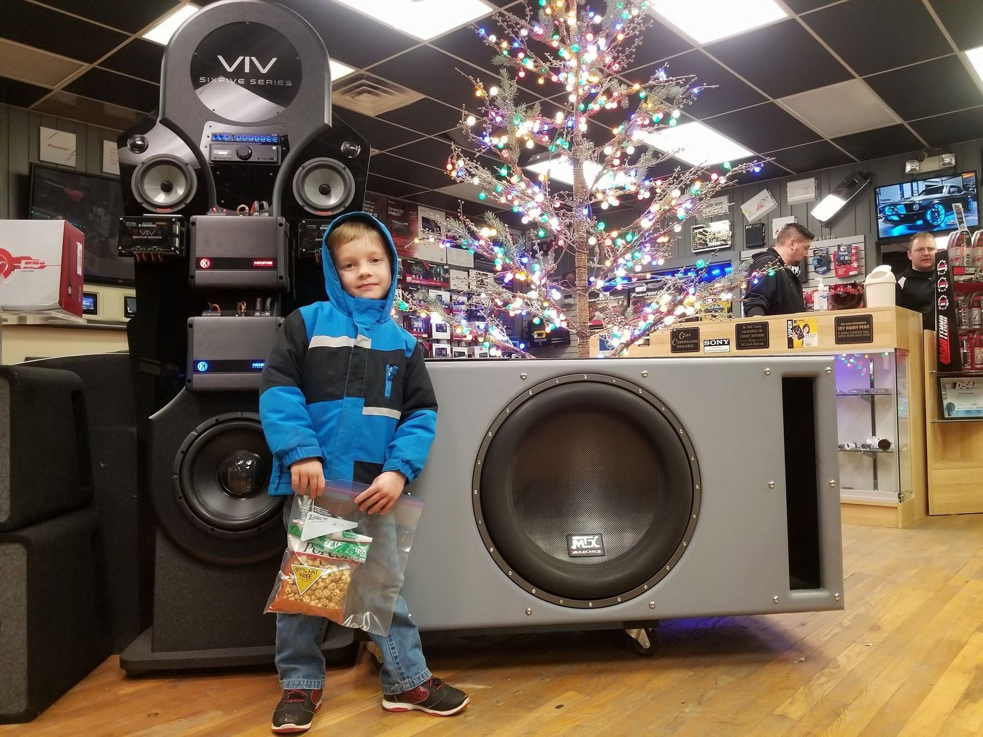 Child standing beside large speakers and subwoofer in a shop display room.