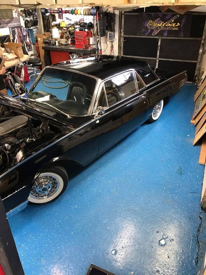 Black classic car in a cluttered garage with blue floor and tools around it