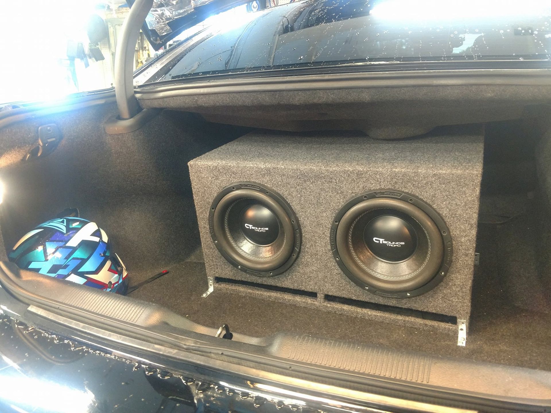 Two black car subwoofers in a gray speaker box installed in a vehicle trunk