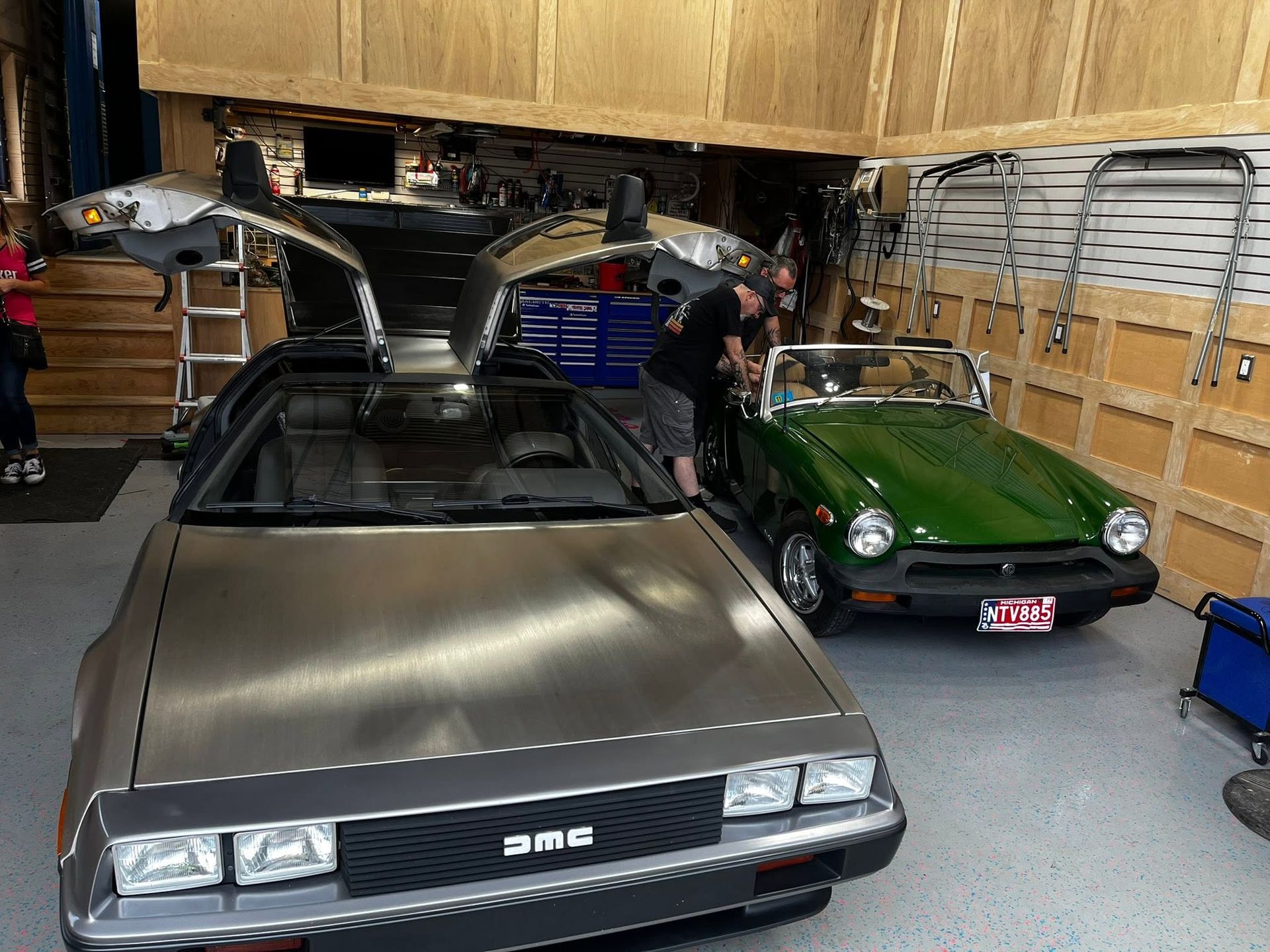 Two DeLorean cars in a garage, one silver with gullwing doors open and one green parked beside it.