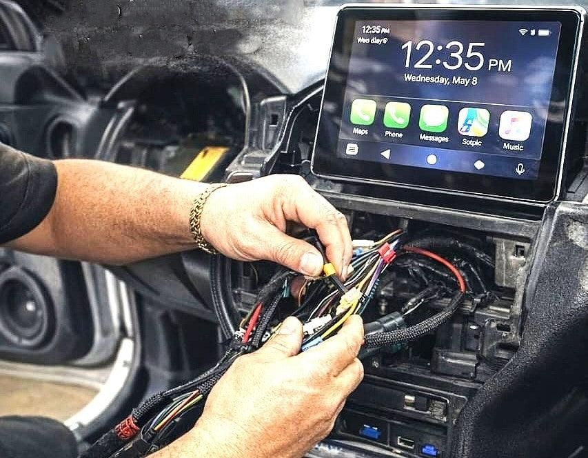 Hands wiring a car dashboard behind a touchscreen infotainment display.