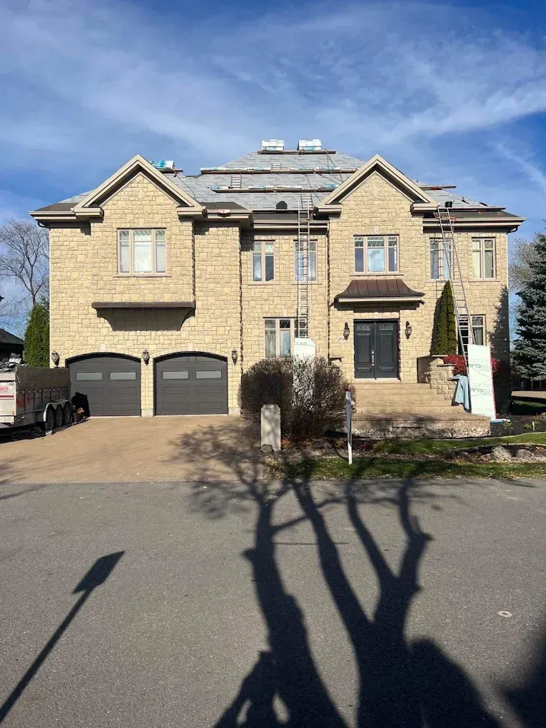 Large modern brick house with a double garage and driveway under a blue sky, tree shadows in front